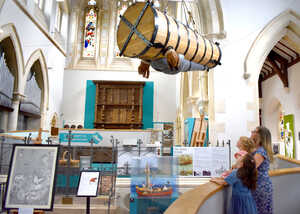 A woman and child observe a large, suspended barrel in a museum setting.
