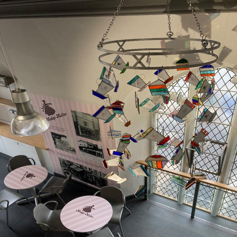An area with tables and chairs. The table has the 'Madge Mellor' logo from the historic cafe on it. Hanging from the ceiling is a mobile made from stained glass nodels of cakes and tea cups