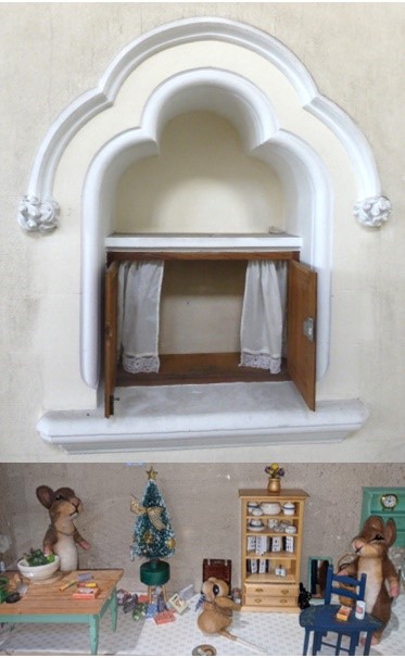 a wooden cupboard in a decorative stone alcove. Inside the cupbord is a family of felt mice celebrating christmas