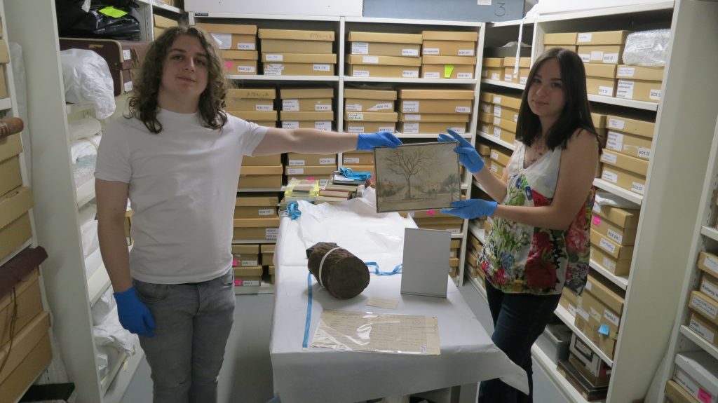 A Man and a Woman wearing blue nitrile gloves holding a watercolour painting of an oak tree. There is a log, Sign and a few documents ahead of them and they are in a store room.