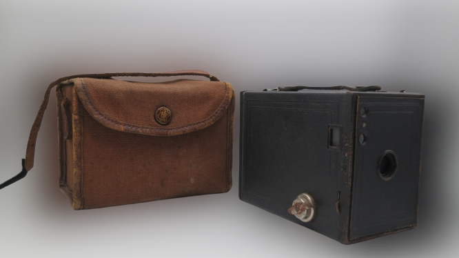 A worn black brownie camera (1920s) with a brown canvas case to the left.