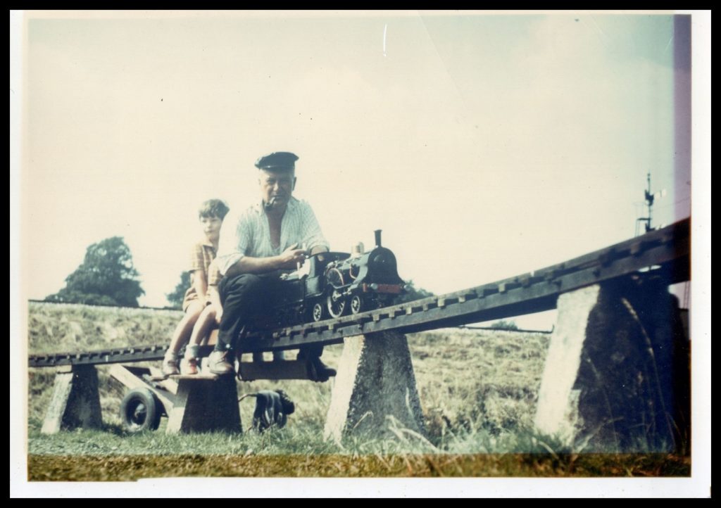 man with pipe driving a model train, two small boys sit behind him